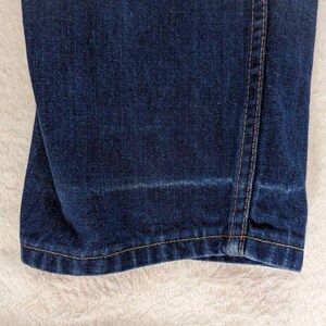 May include: Close-up of dark blue denim jeans. The jeans have a straight leg cut with a light wash detail near the hem. The stitching is a contrasting tan color, and the fabric appears to be slightly worn.