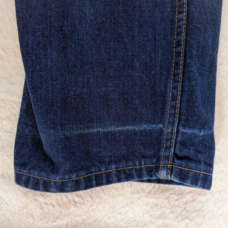 May include: Close-up of dark blue denim jeans. The jeans have a straight leg cut with a light wash detail near the hem. The stitching is a contrasting tan color, and the fabric appears to be slightly worn.