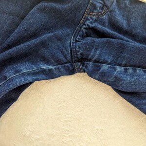 May include: Close-up of dark blue denim jeans. The jeans show the front fly, pockets, and seams with visible stitching. The fabric has a slightly faded appearance, and the jeans are resting on a cream-colored, textured surface.