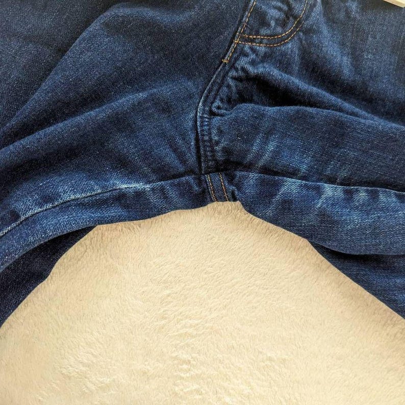 May include: Close-up of dark blue denim jeans. The jeans show the front fly, pockets, and seams with visible stitching. The fabric has a slightly faded appearance, and the jeans are resting on a cream-colored, textured surface.