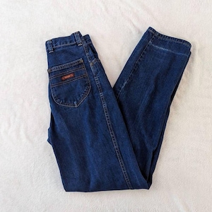 May include: Dark blue denim jeans with a classic five-pocket design. The jeans feature a high waist and a straight leg cut. The Wrangler logo is visible on the back pocket. The jeans are folded neatly on a white surface.