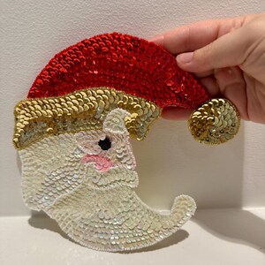 Può includere: Un ornamento natalizio a forma di mezzaluna con un viso di Babbo Natale, realizzato con paillettes bianche, dorate e rosse. L'ornamento ha un cappello rosso con un bordo dorato.