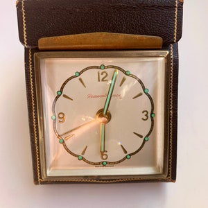 May include: A vintage brown leather-covered alarm clock with a white face and green accents. The clock face reads "Remembrance" and has gold hands and Roman numeral hour markers. The clock is made in Western Germany.