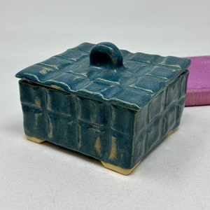 May include: A small, teal blue ceramic box with a woven pattern and a handle on the lid. The box has four small feet on the bottom.