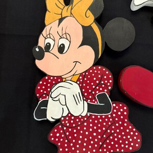 May include: Wooden cutout of Minnie Mouse, featuring a red dress with white polka dots, white gloves, and a yellow bow. The character's face is pink with black eyes and a black nose. The cutout is painted with bright colors.