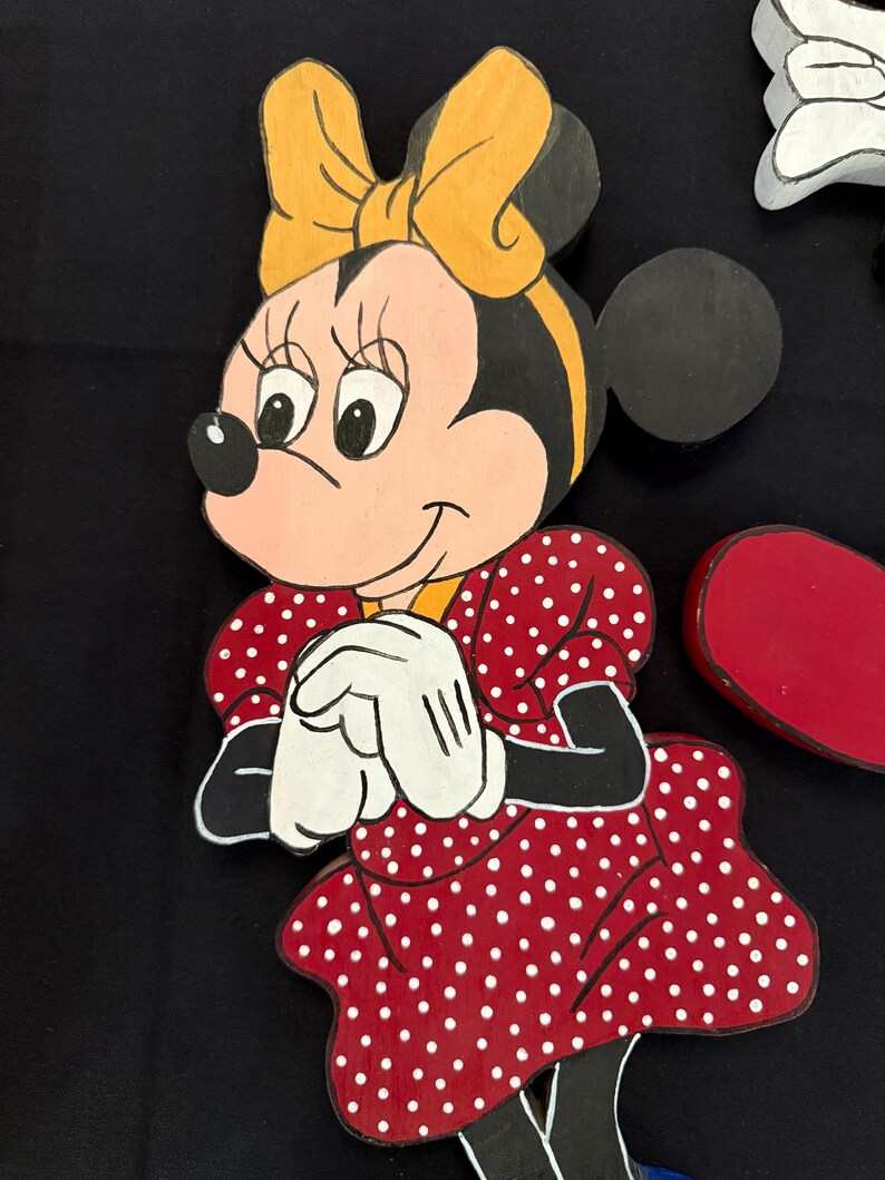 May include: Wooden cutout of Minnie Mouse, featuring a red dress with white polka dots, white gloves, and a yellow bow. The character's face is pink with black eyes and a black nose. The cutout is painted with bright colors.
