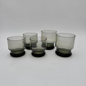 May include: Set of six gray-tinted, lowball glasses. Each glass has a rounded bowl and a dark gray base. The glasses are arranged on a white surface, showcasing their simple, modern design. Ideal for serving cocktails or other beverages.