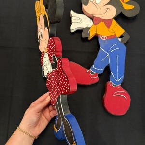 May include: Wooden cutouts of Mickey and Minnie Mouse. Minnie is wearing a red polka dot dress, and Mickey is in blue pants and a yellow shirt. Both have red shoes. The figures are painted with black outlines and details.