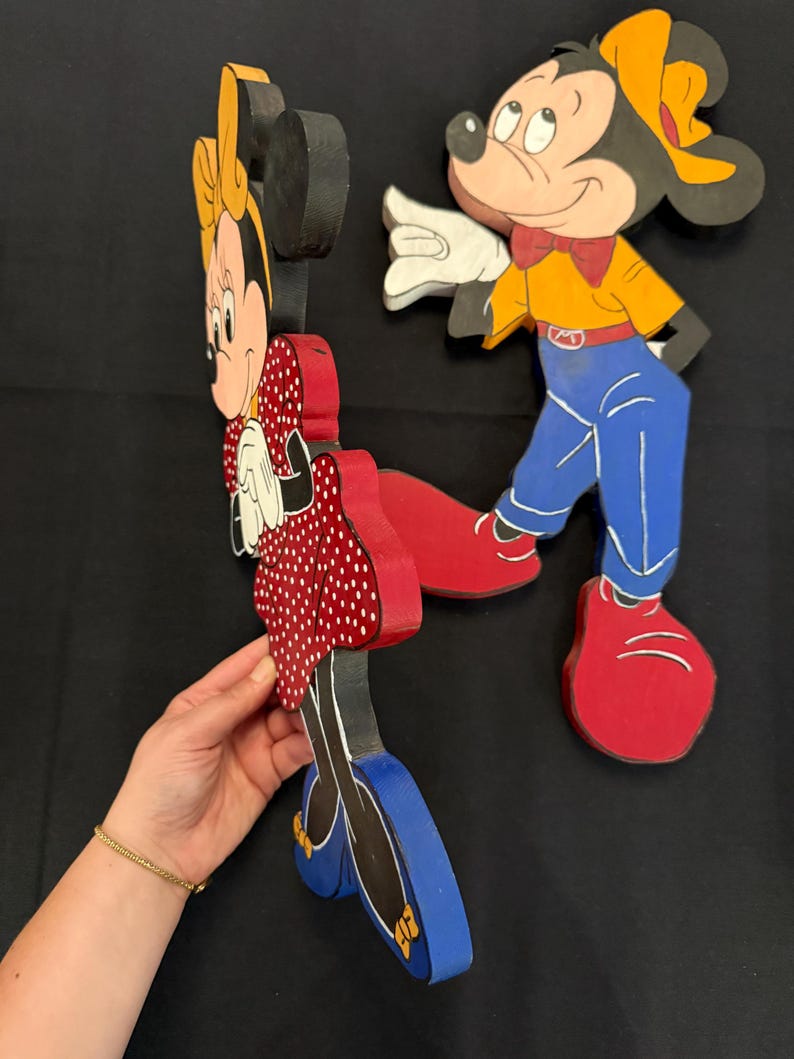 May include: Wooden cutouts of Mickey and Minnie Mouse. Minnie is wearing a red polka dot dress, and Mickey is in blue pants and a yellow shirt. Both have red shoes. The figures are painted with black outlines and details.