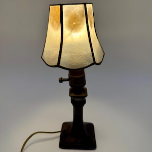 May include: A small, antique table lamp with a square base and a dark brown finish. The lamp has a textured, off-white lampshade with black trim. The lamp is lit, and the cord is visible.
