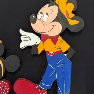 May include: Wooden cutout of Mickey Mouse in a walking pose. Mickey is wearing a yellow shirt, blue pants, red shoes, and a red bow tie. His ears are black, and he has a yellow hat. The cutout is painted with bright colors and has a cartoon style.