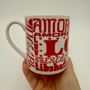 May include: White ceramic mug with red text that says "AMOR" and "LOVE" in a decorative font. The mug has a curved handle and is held in a hand.