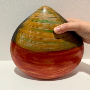 May include: A teardrop-shaped wooden bowl with a multi-colored design. The top half of the bowl is painted in shades of brown, green, and black, while the bottom half is painted in shades of red and orange.
