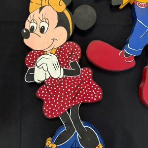 May include: Wooden cutout of Minnie Mouse in a red polka dot dress, yellow bow, and blue shoes. The character is smiling with white gloves and black ears. Part of a larger Disney-themed set.