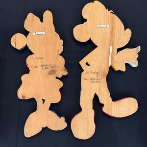 May include: Two wooden wall hangings, one depicting Minnie Mouse and the other Mickey Mouse, with painted details in red, yellow, blue, and black. Each has a metal hanger on the back and handwritten text that reads "To Audrey" and "From Catherine & Kay Helm".