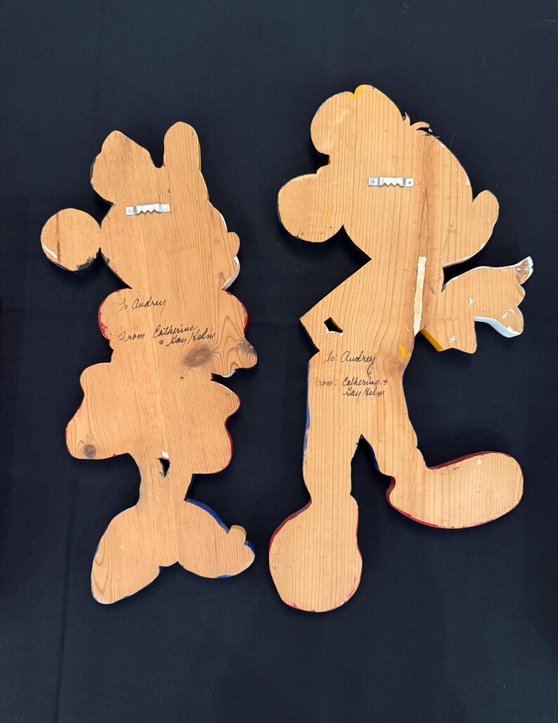 May include: Two wooden wall hangings, one depicting Minnie Mouse and the other Mickey Mouse, with painted details in red, yellow, blue, and black. Each has a metal hanger on the back and handwritten text that reads "To Audrey" and "From Catherine & Kay Helm".