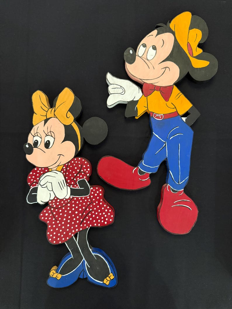 May include: Wooden cutouts of Mickey and Minnie Mouse. Mickey is wearing a yellow shirt, blue pants, red shoes, and a red bow tie. Minnie is wearing a red polka dot dress, blue shoes, and a yellow bow. Both have black ears and are smiling.