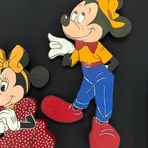 May include: Wooden cutouts of Mickey and Minnie Mouse. Mickey is wearing a yellow shirt, blue pants, red shoes, and a yellow hat. Minnie is wearing a red dress with white polka dots and a yellow bow. Both have white gloves and are smiling.