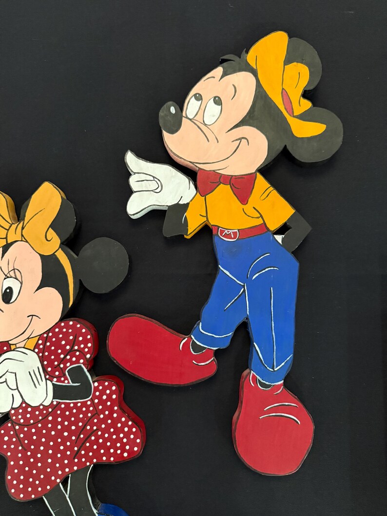 May include: Wooden cutouts of Mickey and Minnie Mouse. Mickey is wearing a yellow shirt, blue pants, red shoes, and a yellow hat. Minnie is wearing a red dress with white polka dots and a yellow bow. Both have white gloves and are smiling.