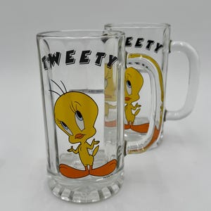 May include: Two clear glass beer mugs featuring the cartoon character Tweety Bird. Each mug has a white handle and a yellow Tweety Bird graphic with the word "Tweety" in black lettering. The mugs are on a white surface.
