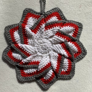 May include: A crocheted pot holder in the shape of a flower with a gray, white, and red color scheme. The pot holder has a loop for hanging.