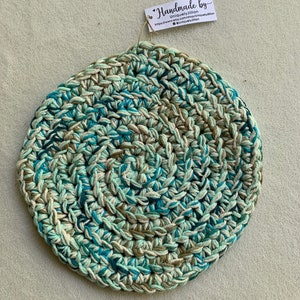 May include: A round crocheted coaster in shades of green, blue, and white. The coaster is handmade and has a tag attached that reads "Handmade by UniquelyJillian".