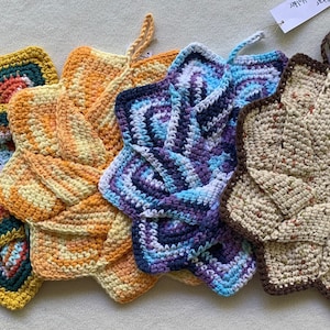 May include: A collection of four handmade star-shaped pot holders in various colors. The pot holders are crocheted with a textured design, featuring shades of yellow, orange, teal, purple, and brown. Each has a loop for hanging.