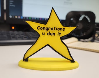 May include: A yellow star-shaped award with a black outline and the text "Congrations u dun it" on it. The star is sitting on a yellow base.