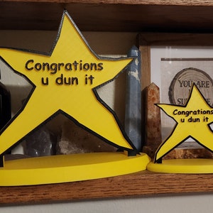 May include: Two yellow star-shaped awards with black outlines and the text "Congratons u dun it" on each. The stars are on a wooden shelf.