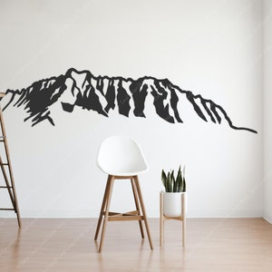 Op de afbeelding: Een zwart silhouet van een bergketen op een witte muur. Een houten ladder en een witte kruk staan op de voorgrond. Een plant in een pot staat aan de rechterkant van de afbeelding.