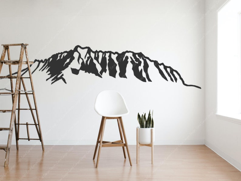 Op de afbeelding: Een zwart silhouet van een bergketen op een witte muur. Een houten ladder en een witte kruk staan op de voorgrond. Een plant in een pot staat aan de rechterkant van de afbeelding.
