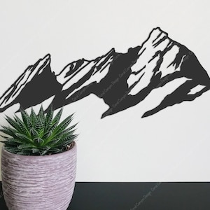 Pu&ograve; includere: Silhouette di metallo nero di una catena montuosa con tre cime. La silhouette &egrave; montata su una parete bianca con una piccola pianta grassa verde in un vaso grigio in primo piano.