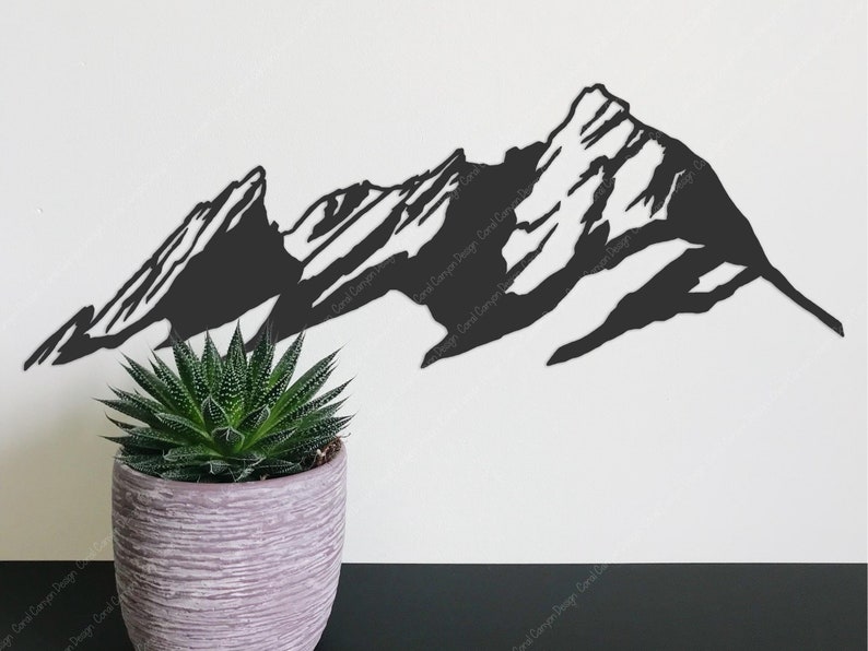 Pu&ograve; includere: Silhouette di metallo nero di una catena montuosa con tre cime. La silhouette &egrave; montata su una parete bianca con una piccola pianta grassa verde in un vaso grigio in primo piano.