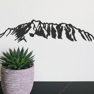 Op de afbeelding: Zwarte silhouet van een bergketen, uit metaal geknipt, tegen een witte muur. Een kleine groene succulent staat in een grijze pot op de voorgrond.