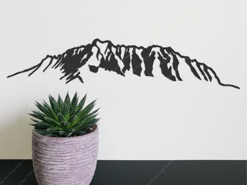 Op de afbeelding: Zwarte silhouet van een bergketen, uit metaal geknipt, tegen een witte muur. Een kleine groene succulent staat in een grijze pot op de voorgrond.
