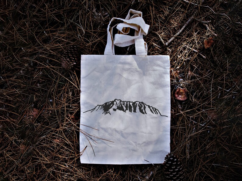 Op de afbeelding: Een witte tote bag met een zwart bergketenontwerp dat op de voorkant is gedrukt. De tas ligt op een bed van bruine dennennaalden.