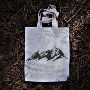 Pu&ograve; includere: Una borsa tote in tela bianca con una silhouette nera di una catena montuosa stampata sul davanti. La borsa &egrave; sdraiata su un letto di aghi di pino marroni.