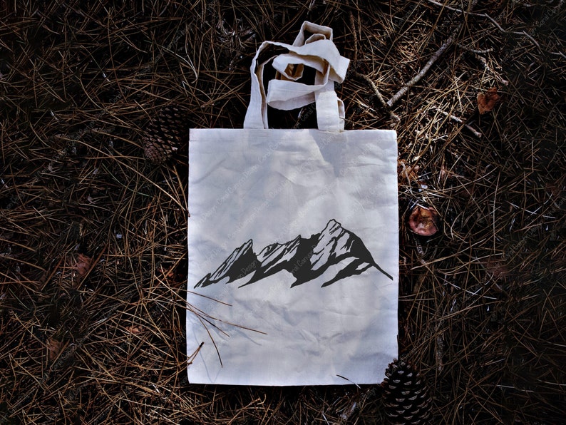 Pu&ograve; includere: Una borsa tote in tela bianca con una silhouette nera di una catena montuosa stampata sul davanti. La borsa &egrave; sdraiata su un letto di aghi di pino marroni.