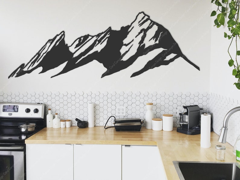 Pu&ograve; includere: Decorazione da parete in metallo nero a forma di catena montuosa in un ambiente da cucina moderno. La decorazione &egrave; montata su una parete bianca sopra un bancone da cucina con armadietti bianchi e una stufa in acciaio inox.