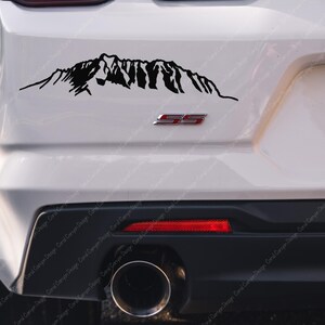 Op de afbeelding: Een zwarte sticker met een silhouet van een bergketen op de achterkant van een witte auto. De auto heeft een zwarte bumper en een rood "SS"-embleem.