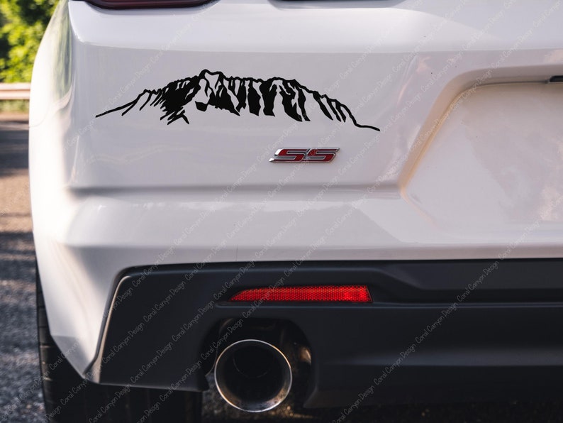 Op de afbeelding: Een zwarte sticker met een silhouet van een bergketen op de achterkant van een witte auto. De auto heeft een zwarte bumper en een rood "SS"-embleem.