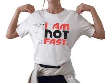 May include: A white short-sleeved t-shirt featuring a black line-art graphic of a round robot character and the red and black text "I AM NOT FAST." The shirt is paired with black athletic shorts and a beige long-sleeved sweatshirt tied around the waist.