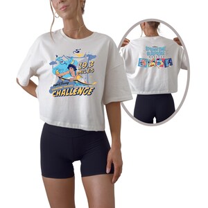 May include: White cropped t-shirt with a graphic featuring Aladdin, Genie, and the text "19.3 Miles Challenge". The back of the shirt displays the text "Springtime Surprise Weekend" and character images. The shirt is paired with black shorts.