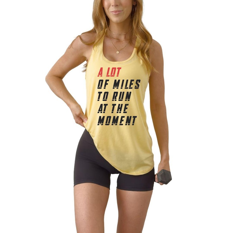 May include: Yellow tank top with the text "A LOT OF MILES TO RUN AT THE MOMENT" in black and red. The person is wearing black shorts and holding a dumbbell.