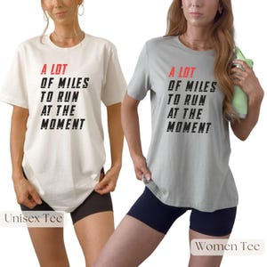 May include: Two t-shirts, one cream and one gray, each with the text "A LOT OF MILES TO RUN AT THE MOMENT" in black and red. The cream shirt is labeled "Unisex Tee" and the gray shirt is labeled "Women Tee". Both shirts are paired with black shorts.