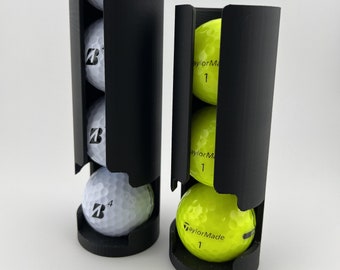 May include: Two black golf ball holders, one tall and one short, each containing golf balls. The tall holder has four white Bridgestone golf balls. The shorter holder has three bright yellow TaylorMade golf balls.