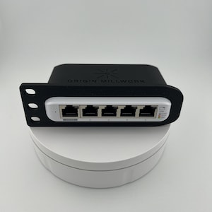 以下が含まれることがあります： 白い前面と各ポートのLEDインジケーターが付いた、黒い5ポートネットワークスイッチ。スイッチには「ORIGIN MILLWORK」というラベルが貼られており、星型のロゴがあります。