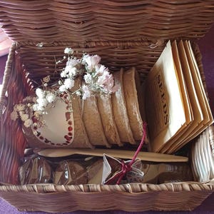 Peut inclure: Un panier en osier rempli de fournitures de jardinage, y compris des sachets de graines, un petit pot et un sac de matière brune. Le panier est doublé d'un tissu blanc et a une poignée brune. Le panier est posé sur une surface violette.