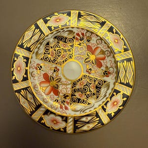 Puede incluir: Un pequeño plato redondo de porcelana con un patrón floral colorido en tonos rojos, azules y dorados. El plato tiene un centro blanco y un borde dorado.