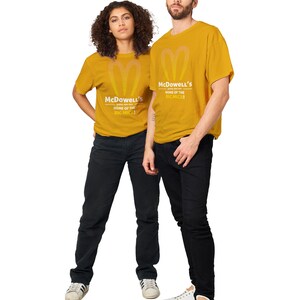 May include: Two people wearing mustard yellow t-shirts with the text "McDowell's HOME OF THE BIG MICK!" and a heart-shaped logo. They are also wearing black jeans and white sneakers. The background is white.